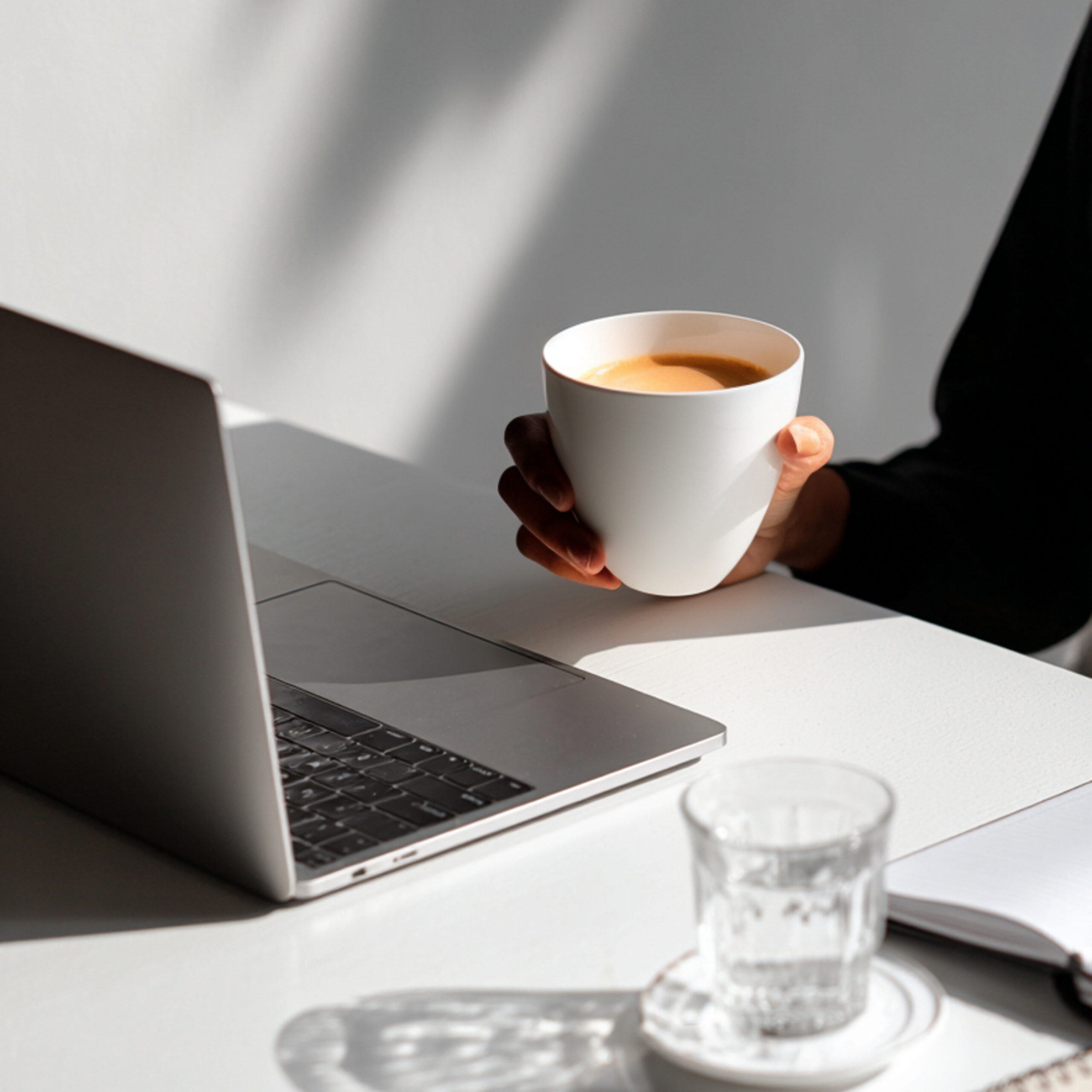 Close-up van een tafel met een opengeklapte laptop, een kop koffie, een glas water en een notitieboekje in de hoek van de foto | Elena & Company