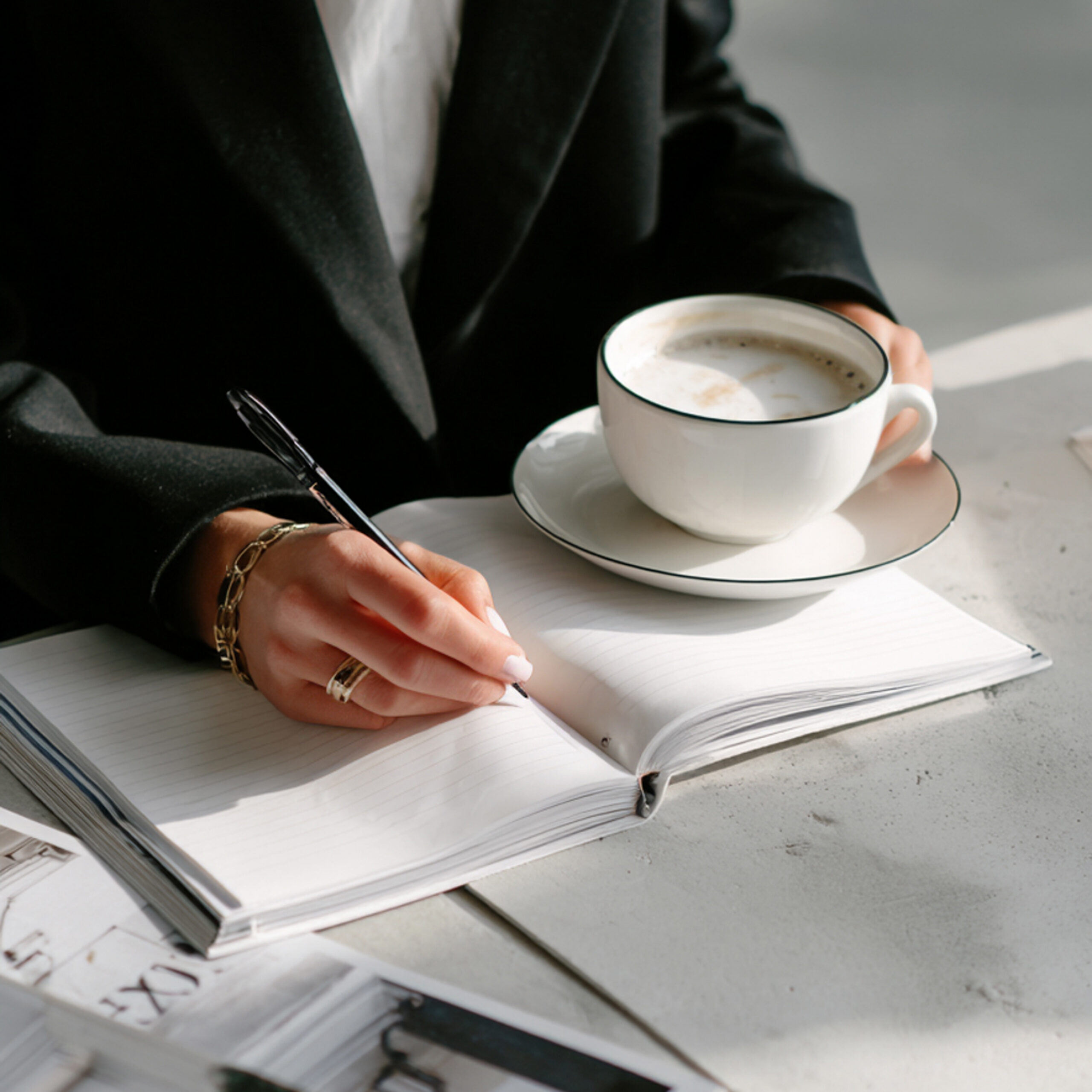 Close-up van een business woman in een zwarte blazer en goudkleurige juwelen. Ze zit aan tafel terwijl ze in haar notitieboek schrijft en haar capuccino in haar ander hand houdt | Elena & Company