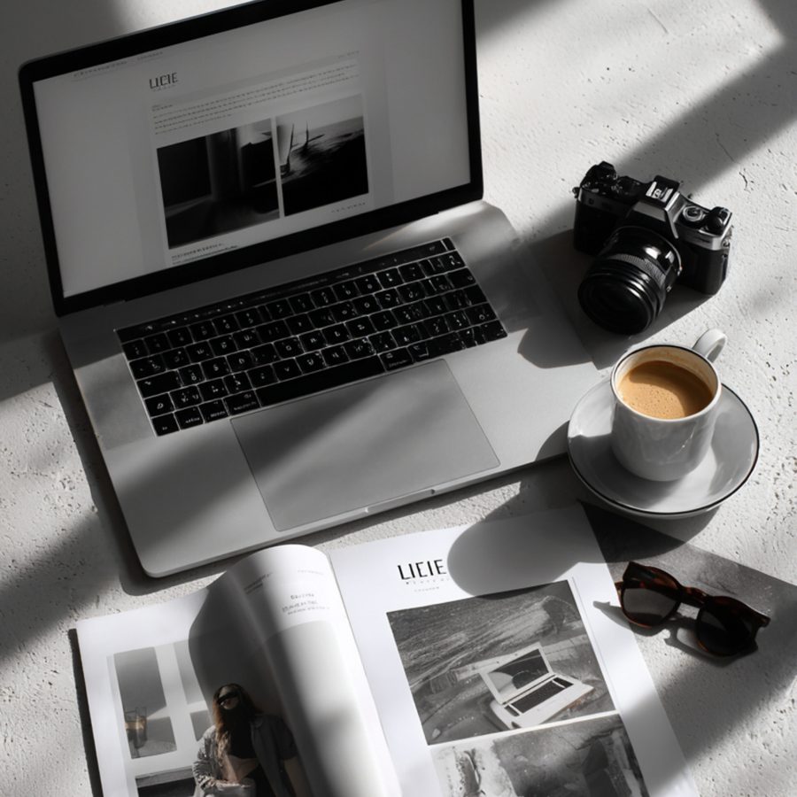 Opengeklapte laptop op een witte marmeren tafel, er omheen liggen opengeslagen tijdschriften met mooie zwart-wit foto's, een zwarte zonnebril, een kop koffie op een schoteltje en een zwarte systeemcamera, de essentiële tools van een business woman | Elena & Company